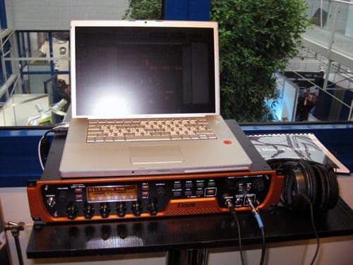 A laptop positioned on top of a black and orange audio interface, showcasing various knobs and inputs. A pair of headphones is connected to the interface. In the background, a green indoor plant and some blurred structures are visible.