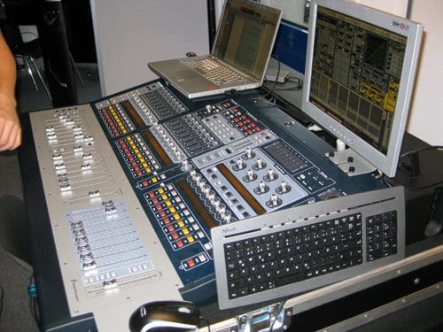 A sound mixing console with various knobs, sliders, and buttons, alongside two computer monitors displaying audio software, all set on a portable rack. A hand is partially visible on the left side, interacting with the equipment.