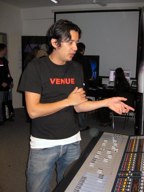 A man wearing a black t-shirt with the word 'VENUE' in red, gesturing while discussing equipment at a sound mixing station. In the background, other individuals are working on computers in a well-lit room.