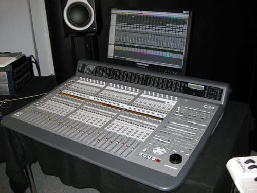 A gray digital audio mixing console with numerous sliders and buttons, prominently displayed on a black table. A monitor above shows a colorful audio editing interface, while a studio monitor speaker is visible beside the console.