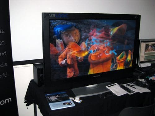 A large flat-screen television displaying animated characters from a film, featuring vibrant colors and playful designs. The setup includes a black table with various printed materials and electronic equipment, set against a dark background.