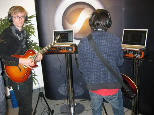 Two musicians engaged in a live music demo in a modern space. One musician plays an electric guitar, while the other interacts with a laptop setup. Headphones are worn by both individuals, and there are decorative plants in the background.