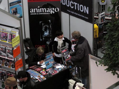 A busy exhibition scene featuring a woman at a booth displaying colorful magazines and merchandise. She is engaged in conversation with a man, while another person is seated, reviewing materials. Surrounding banners advertise the animago award and digital production.