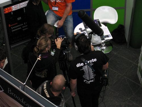 A group of three individuals, focused on operating a video camera, are gathered around a filming setup. One person is wearing a black shirt with white text, and another is seated on a modern white stool. The space is vibrant with colorful walls and broadcasting equipment.