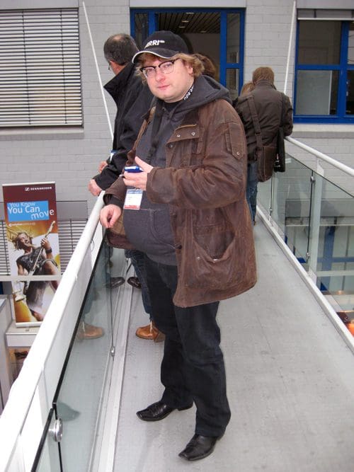 A man with glasses and curly hair stands on a glass walkway, wearing a brown jacket and black pants. He has a slight smile and is gesturing with one hand. In the background, other attendees are partially visible.