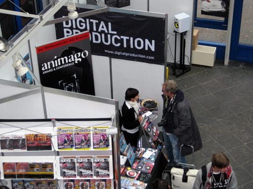 An overhead view of a trade show booth with a large sign reading 'DIGITAL PRODUCTION'. A woman in a black and white outfit talks to a man holding a camera at the booth, surrounded by promotional materials and magazines.