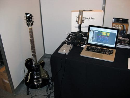 An electric guitar stands next to a microphone and a laptop on a black table. The laptop screen displays music production software, and headphones are resting nearby. The background is a simple, neutral setting.