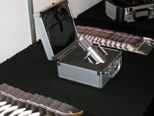A silver microphone placed inside a protective case on a black table. The case is slightly open, revealing the microphone nestled in foam padding. In the background, a textured surface extends across the table.