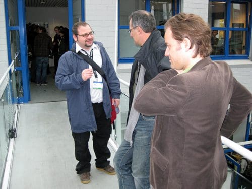 Three men engaged in conversation on a balcony. One man wears glasses and a blue jacket, another has short gray hair and a black coat, while the third has brown hair and wears a brown blazer. The background includes a building with blue windows.