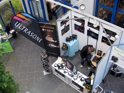 An aerial view of an exhibition booth featuring ULTRASONE branding, with a variety of headphones displayed on a table. A person is working on a laptop at the booth, surrounded by promotional materials and posters.