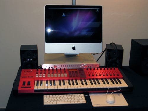 A sleek workspace featuring an iMac computer with a vibrant screensaver, an electronic keyboard filled with colorful keys, and two black studio monitors on either side. A white keyboard and mouse are placed on a black table surface.