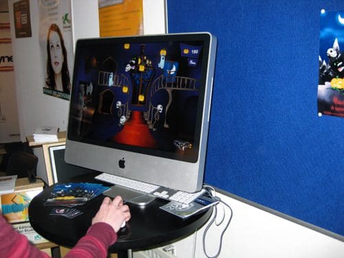A person using an Apple iMac computer to play a colorful cartoon-style game featuring a spooky castle scene with animated characters. The desk is cluttered with brochures and posters on the wall behind.