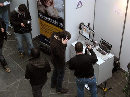 A group of people interacting at a tech exhibition, with one individual wearing headphones while examining a setup that includes a laptop and a musical instrument. In the background, a banner displays the name "Das Medien-Studio."