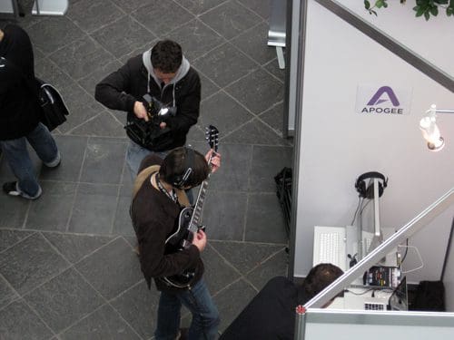 An overhead view of a music event, featuring a man playing an electric guitar while wearing headphones. Another man is filming him with a camera. In the background, a table with equipment and a sign for Apogee is visible.