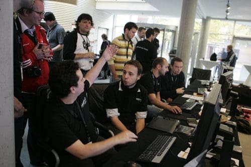 A group of people gathered in a modern venue, with several individuals working on computers at a table. Some attendees engage in conversation, while others focus on their screens, contributing to a collaborative atmosphere.