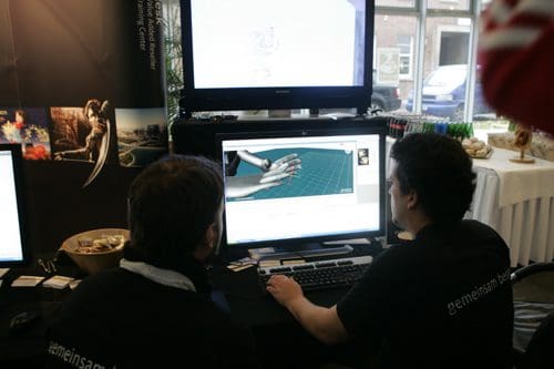 Two individuals focused on a computer screen displaying 3D modeling software, with a large monitor showing graphics in the background. The setting appears to be a tech exhibition, with a table featuring snacks and drinks.