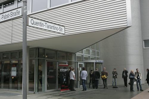 A group of people standing in line outside a modern building with a silver exterior. A street sign reads 'Quentin-Tarantino-Straße.' The entrance features large glass doors and a promotional banner visible.