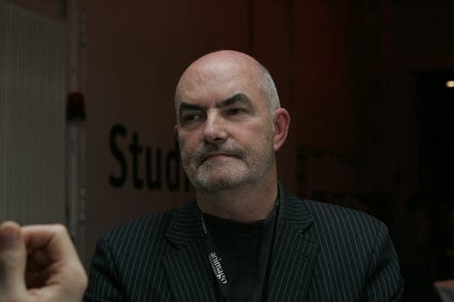 A middle-aged man with a beard, wearing a pinstripe blazer and a black shirt, sits at a table with an expressive facial expression. The background features muted lighting and text that reads 'Studio'.