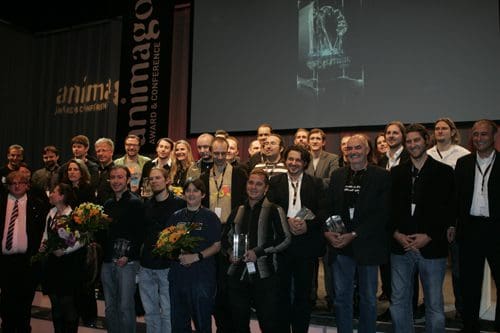 A large group of people standing on stage at an awards ceremony, posing for a photo. They hold trophies and flowers, with banners in the background promoting the event. The atmosphere is celebratory.