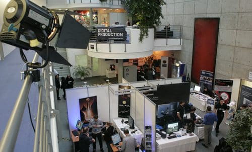 A wide-angle view of an exhibition hall featuring various booths related to digital production. Attendees interact with displays and products, while a spotlight illuminates the area from above. Greenery is visible near the upper level.
