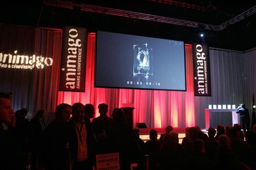 A large stage at an event featuring illuminated banners with the word 'animago.' A countdown timer is displayed on a screen, while an audience of dark silhouettes watches attentively. Soft red lighting enhances the atmosphere.