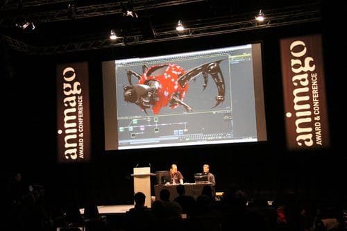 A stage setting with two presenters at a table, demonstrating a digital animation program on a large screen showing a detailed, animated character design. On both sides, banners display the event name 'animago'.