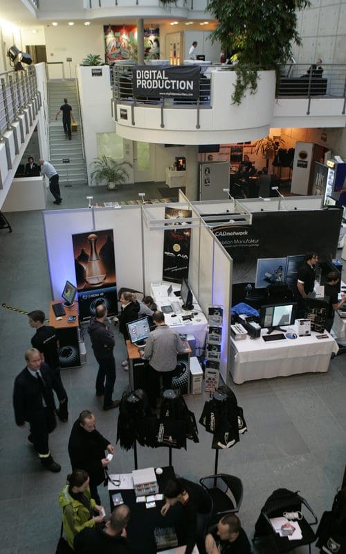 A spacious venue featuring a digital production exhibition. Attendees engage at various booths displaying screens and promotional materials. A large sign overhead reads 'DIGITAL PRODUCTION,' with a staircase visible nearby.