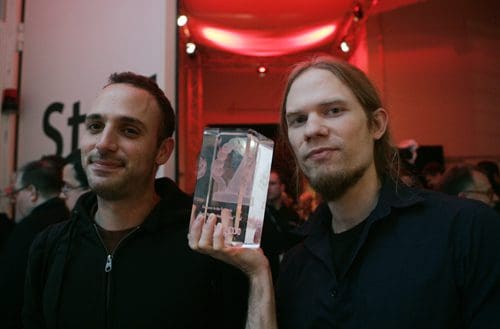 Two men are smiling in a crowd, one holding a clear award trophy in front of him. The background is filled with blurred figures and a warm red light enhances the celebratory atmosphere.