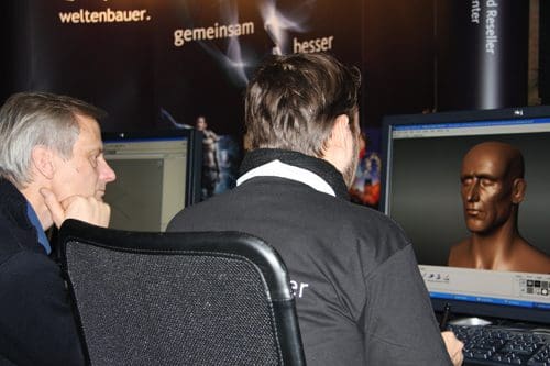 Two individuals sit at computer desks, focused on a digital art program displaying a 3D modeling of a human head. The background features promotional banners and colorful graphics.