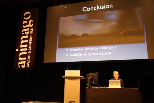 A speaker presenting at the animago Award & Conference, with a slide showing the conclusion of a presentation on animation-centric proceduralism and scalability vs. quality tradeoff, in a darkened auditorium.