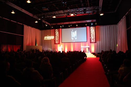 An audience seated in a large, dimly lit room at a conference event, facing a stage with a large screen displaying presentations. Red lighting enhances the atmosphere, and banners featuring the word 'animago' are prominently displayed.