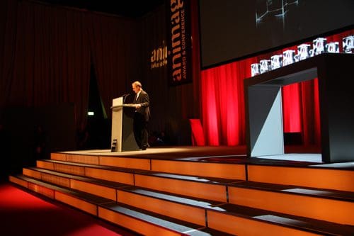 A speaker delivering a presentation at an awards ceremony on a stage with bright red curtains. The podium is in the foreground, and several illuminated awards are displayed on a table behind. Soft lighting creates a formal ambiance.