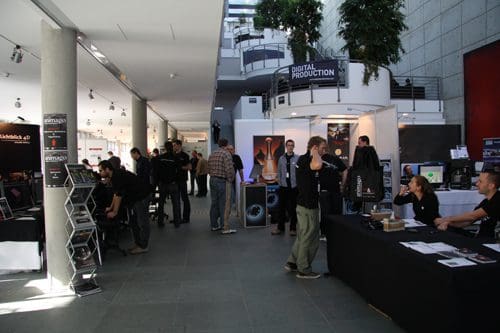 A busy exhibition hall featuring booths for digital production. Attendees engage in discussions, and several people are seen networking. Tall plants are situated throughout the space, creating a welcoming atmosphere.
