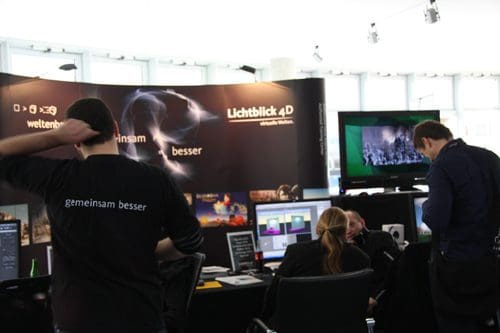 A trade show booth featuring the company name "Lichtblick 4D" with a dark backdrop displaying images. Several people are engaged at workstations with computer screens, while a monitor showcases a video presentation.