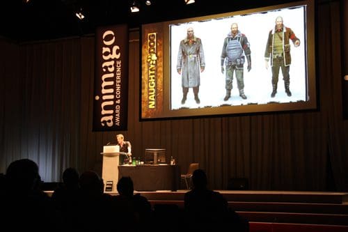 A presenter stands at a podium during a conference, with a large screen displaying character designs from Naughty Dog's game. Audiences in dark seats watch attentively. The screen features three distinct character designs.