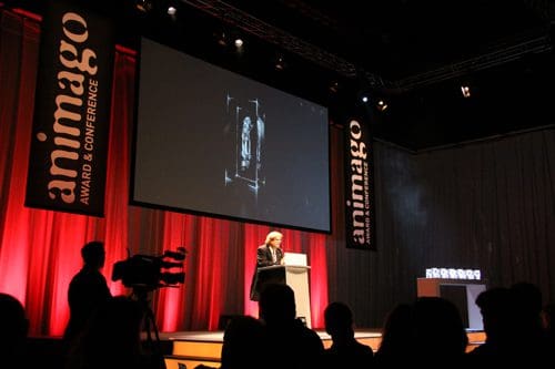 A speaker at a podium during the animago awards and conference event, with a large screen displaying an animated figure. The stage is lit with red lighting, and several award trophies are visible on a table at the side.