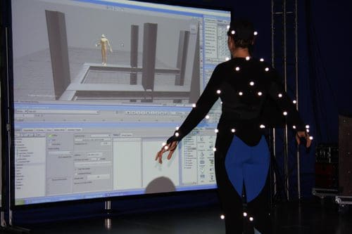 A person wearing a black motion capture suit with white markers stands in front of a large screen displaying a 3D animation of a figure navigating through a digital environment. The scene includes a control panel on the screen.