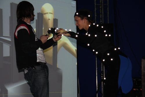 A male and female performer on stage during a motion capture demonstration. The female is wearing a black suit adorned with tracking lights. A screen in the background shows a digital figure in action.