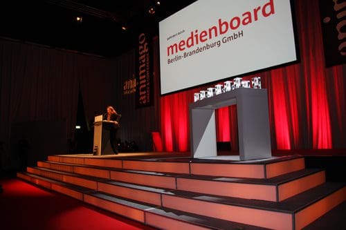 A speaker stands at a podium on a large stage during an event, with a backdrop displaying 'medienboard Berlin-Brandenburg GmbH'. The stage is illuminated with red and white lighting, and several awards are arranged on a platform.