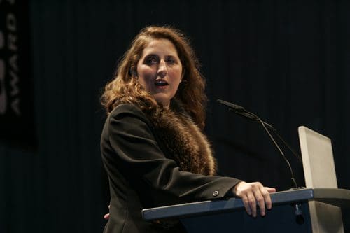 A woman with long, wavy brown hair stands at a podium, delivering a speech. She wears a black jacket with a fur collar, focusing intently on her audience. The background is dark, emphasizing her presence at the event.
