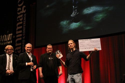 Five men on stage at an awards ceremony. One man holds a trophy and a large check, smiling, while others stand beside him, wearing formal attire. A large screen displays visuals in the background, and red curtains frame the stage.