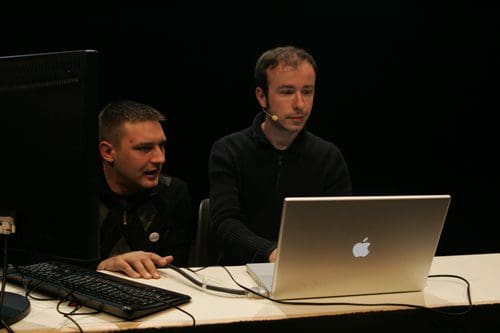 Two men at a white table, one is using a laptop while the other appears to be consulting him. A black screen is visible in the background, and a keyboard is placed in front of them.