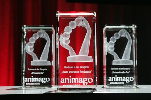 Three glass awards with etched designs of raised fists, displayed on a table. The awards are labeled 'animago AWARD 2009', recognizing achievements in categories such as 'Best Interactive Production'.