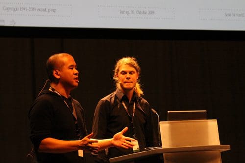 Two speakers stand at a podium during a presentation. One man, with short dark hair, gestures while speaking, wearing a black shirt. The other, with long light hair, listens attentively, dressed in a black outfit. A presentation screen is slightly visible in the background.