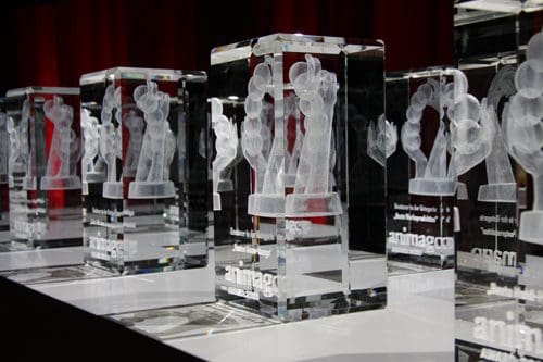 A row of crystal awards displayed on a table, showcasing intricate frosted designs of figures within each transparent block. The awards are illuminated against a backdrop of red curtains.