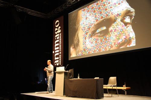 A speaker presenting at a conference on stage with a large projection displaying a close-up of a face adorned with colorful dots. The setting includes a table with chairs and a bottle of water.