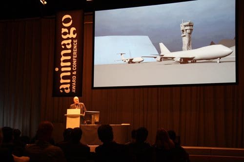 A speaker presents at the animago Award and Conference, with a large projection of a 3D model of an aircraft and a control tower behind them. An audience attentively listens in a dimly lit auditorium.