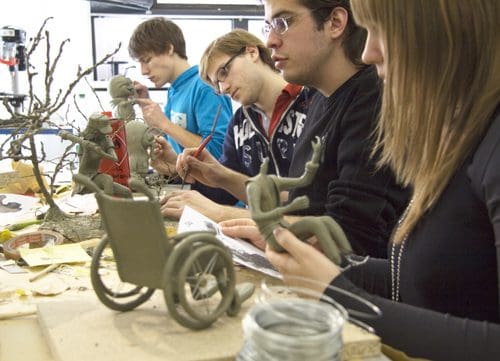 Four students engaged in a sculpting workshop, focusing on creating clay figures. The table is cluttered with various materials, including a wheelchair sculpture and tools for modeling. Each student is intently shaping their artwork.