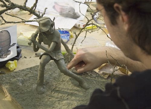 An artist carefully sculpting a clay figure of a warrior, holding a weapon, at a workspace cluttered with tools and reference images. Twigs are visible in the background, adding to the creative atmosphere.