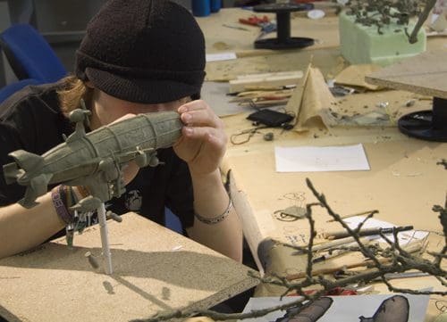A person wearing a black beanie closely examines a model zeppelin while working at a cluttered table filled with crafting materials, tools, and several small sculptures.